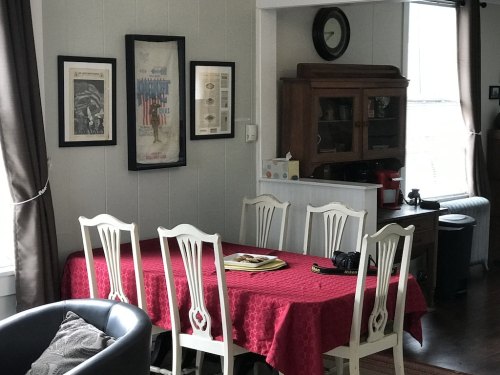 A dining area with a red tablecloth, white chairs, framed art on the walls, and a wooden cabinet in the background.