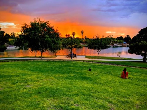 A serene park scene at sunset, featuring a lake, trees, and a person sitting on the grass, enjoying the view.