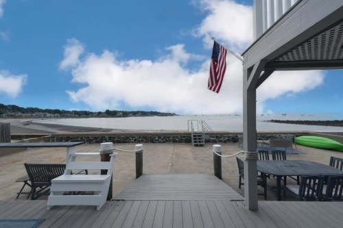 View of a beach with a wooden deck, American flag, and calm water under a blue sky with clouds.