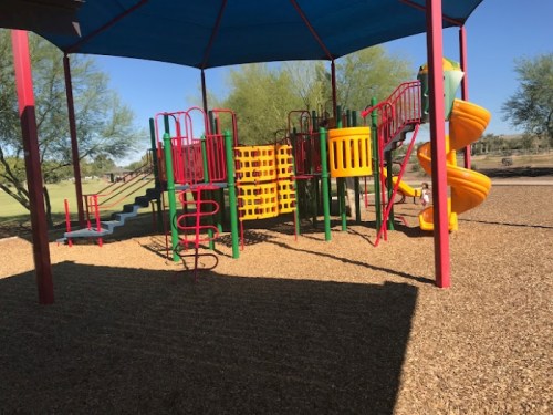 Colorful playground equipment with slides and climbing structures under a shaded canopy, surrounded by gravel.