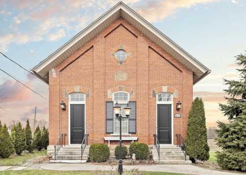 A brick building with a gabled roof, two front doors, and landscaped greenery under a colorful sky.