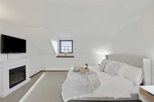 Cozy bedroom with a gray tufted bed, a TV on the wall, and a fireplace, featuring soft lighting and a neutral color palette.