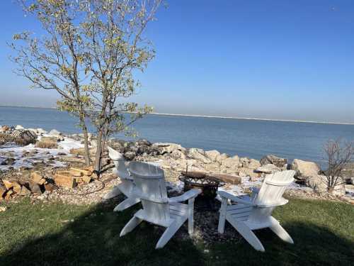 A serene lakeside view featuring white Adirondack chairs, a small fire pit, and a tree against a clear blue sky.