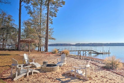 A serene lakeside scene with white chairs around a fire pit, sandy beach, and a dock under a clear blue sky.