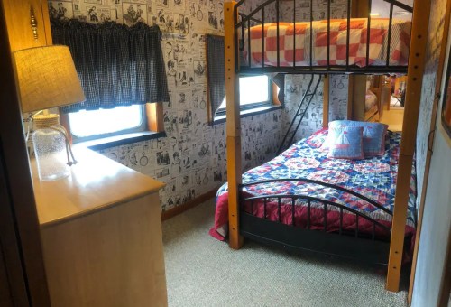 Cozy bedroom with a bunk bed, a single bed, patterned quilts, and a lamp beside a wooden dresser.