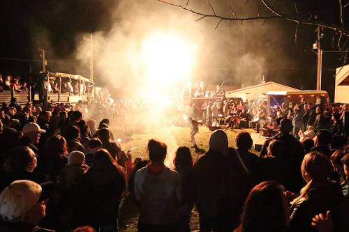 A large crowd gathers around a bright bonfire at night, with smoke rising and people enjoying the festive atmosphere.
