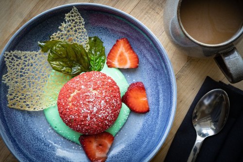 A vibrant dessert featuring a red cake on green cream, garnished with strawberries, basil leaves, and a lace cookie, beside coffee.