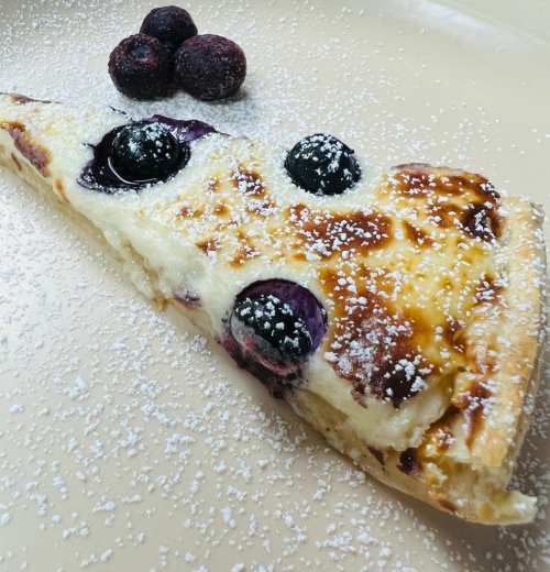A slice of creamy blueberry cheesecake topped with powdered sugar and three blueberries on the side.