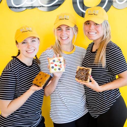 Three smiling women in striped shirts and yellow hats hold decorated cookies against a bright yellow background.
