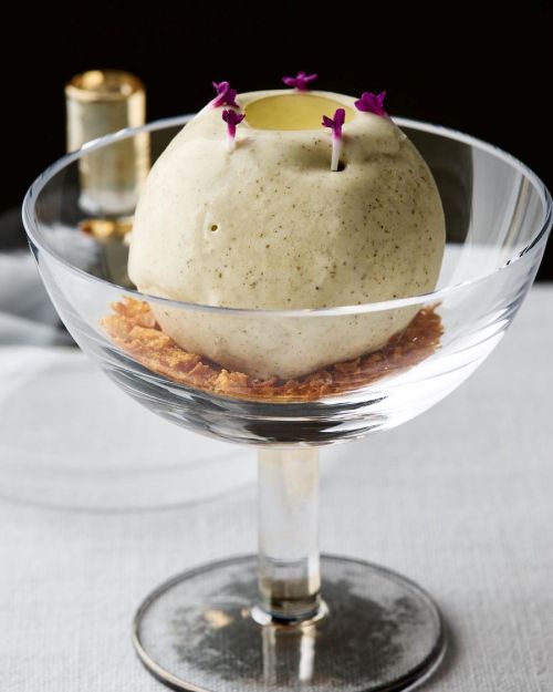 A spherical dessert topped with edible flowers, resting on a crispy base in a clear glass dish.