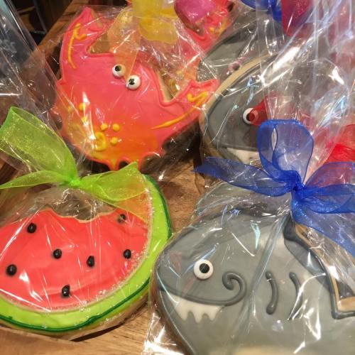 Colorful decorated cookies shaped like a crab, watermelon, and shark, wrapped in clear plastic with colorful ribbons.