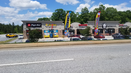 A row of businesses including Pizza Hut, with colorful banners and cars parked in front on a sunny day.