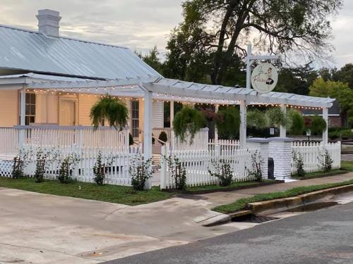 A charming white house with a metal roof, surrounded by greenery and string lights, featuring a sign on the porch.