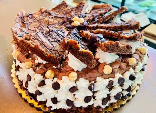 A decadent chocolate cake topped with whipped cream, chocolate drizzle, and cookie pieces, garnished with hazelnuts.