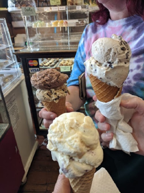 Three ice cream cones in hands, featuring chocolate, cookies and cream, and vanilla flavors, in a colorful ice cream shop.