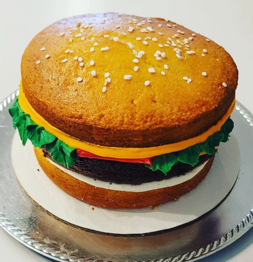 A cake designed to look like a hamburger, featuring layers of yellow cake, chocolate cake, and colorful frosting toppings.