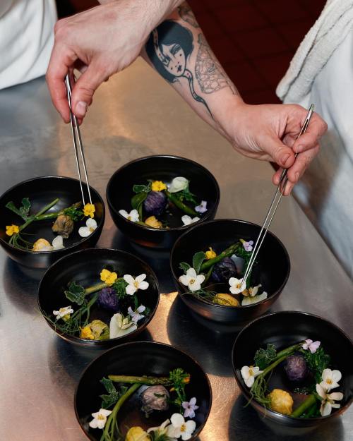 A hand with chopsticks garnishes bowls of colorful food, decorated with flowers and herbs, on a stainless steel surface.