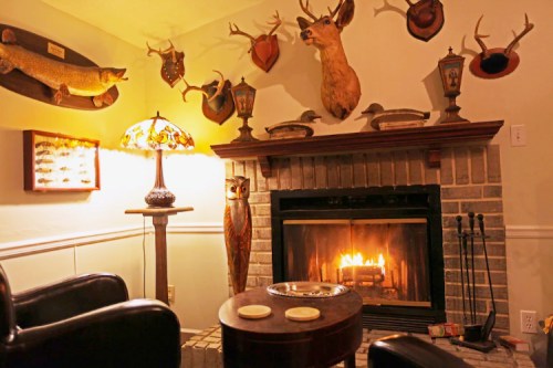 Cozy room with a fireplace, mounted animal heads, a lamp, and a small table with plates, creating a rustic atmosphere.