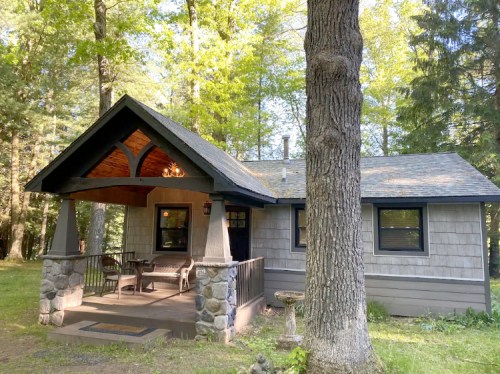 A cozy cabin surrounded by trees, featuring a porch with stone pillars and a sloped roof.