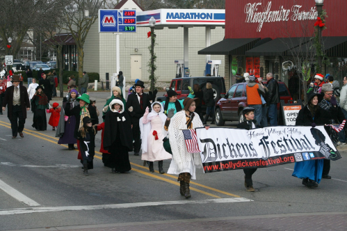 A festive parade with participants in Victorian costumes holding a banner for the Dickens Festival.