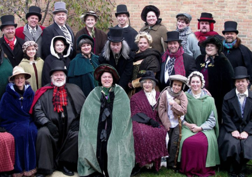 A large group of people dressed in Victorian-era clothing, posing together outdoors with hats and capes.