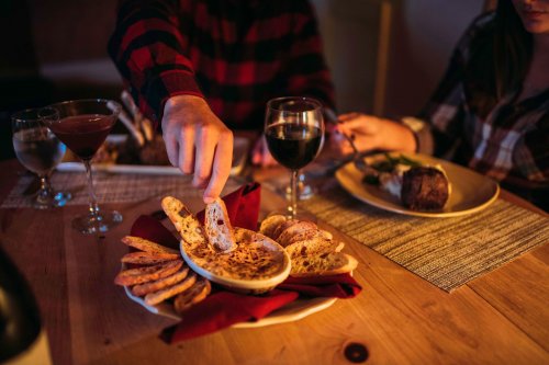 A cozy dinner scene with a person dipping bread into a dish, surrounded by wine glasses and a plated meal.