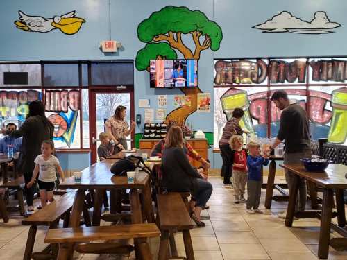 A colorful indoor play area with families, children, and tables, featuring a mural of a tree and a TV on the wall.