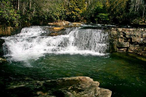 A serene waterfall cascades over rocky ledges into a clear pool, surrounded by lush greenery and trees.