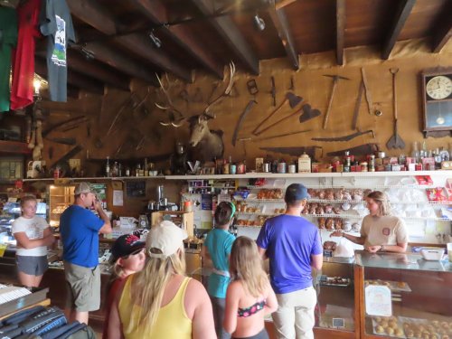 A rustic shop interior with a mounted deer head, shelves of snacks, and customers interacting with the staff.