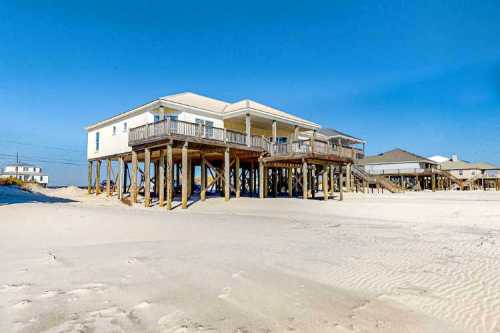 A beach house on stilts overlooking a sandy shore under a clear blue sky.