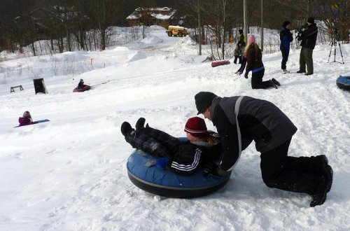 A child on a snow tube is pushed down a snowy hill, while others enjoy tubing in the background.