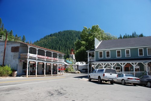A small town street with shops, a white building, and parked cars, surrounded by green hills under a clear blue sky.