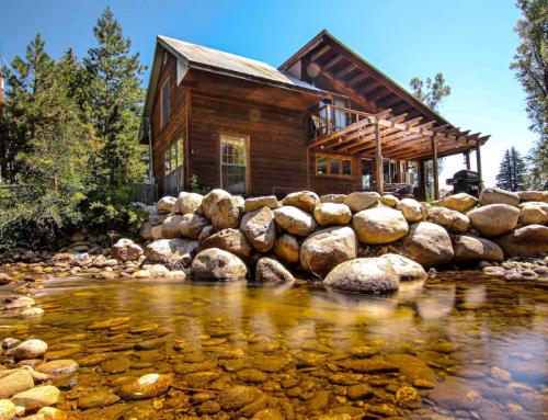 A rustic wooden cabin by a clear stream, surrounded by trees and rocks under a bright blue sky.