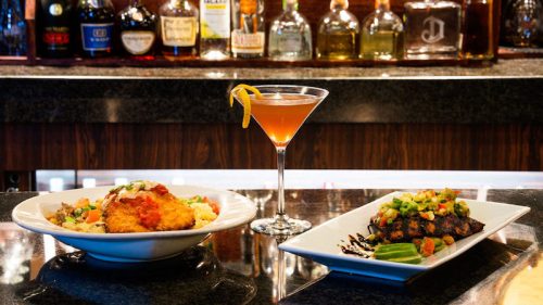 A bar scene featuring two gourmet dishes and a cocktail on a dark countertop, with bottles in the background.
