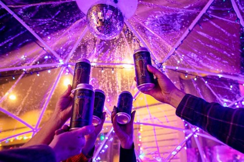 Hands holding cans of beer toast under a disco ball, surrounded by colorful lights and a festive atmosphere.
