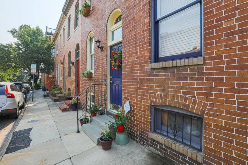 A charming brick building with a blue door, flower pots, and a small staircase, set on a sunny street.