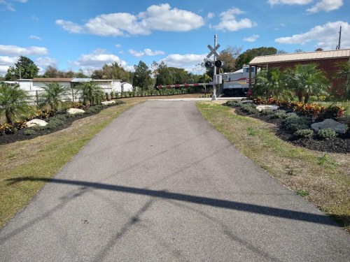 A paved path leads to a railroad crossing, flanked by landscaped gardens and blue skies with scattered clouds.