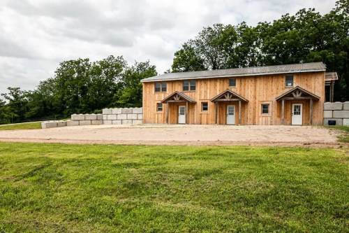 A wooden building with three entrances, surrounded by grassy land and concrete blocks, under a cloudy sky.
