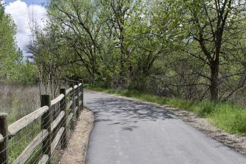 A winding path through a lush green landscape, bordered by a wooden fence and trees under a partly cloudy sky.