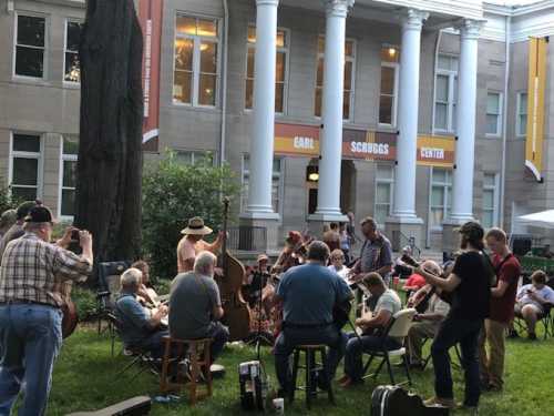 A group of musicians playing instruments outdoors in front of a building, with people seated on chairs and the grass.