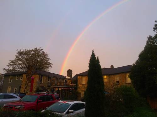 A double rainbow arcs over a residential building with trees and parked cars in the foreground.