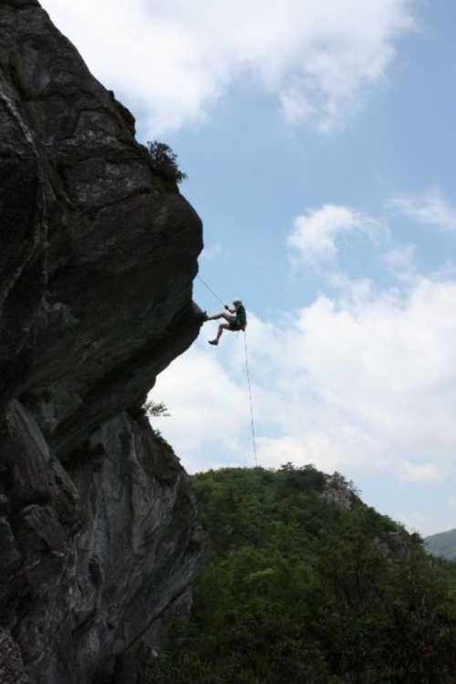 A climber rappels down a rocky cliff against a backdrop of green hills and a cloudy sky.