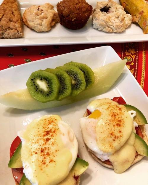 A plate with two eggs Benedict, sliced kiwi, cantaloupe, and assorted baked goods in the background.