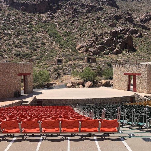 An outdoor amphitheater with red seats, surrounded by rocky hills and greenery, under a clear blue sky.