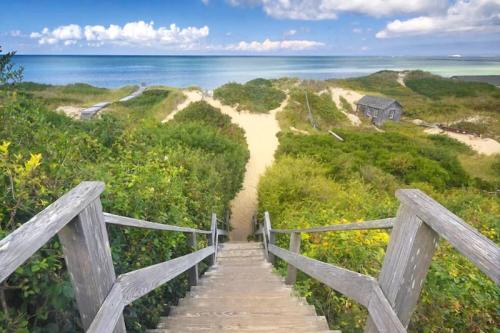 Wooden stairs lead down to a sandy beach surrounded by lush greenery and a calm ocean under a partly cloudy sky.