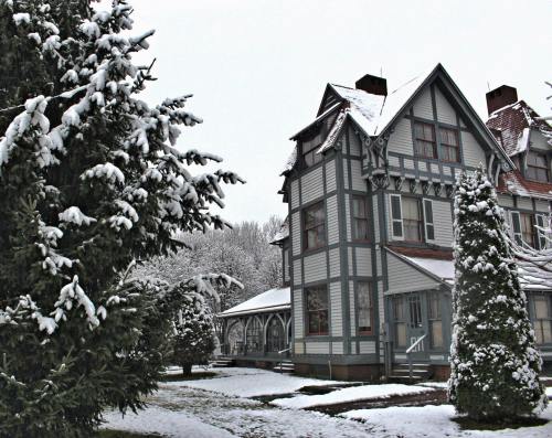 A snow-covered Victorian house surrounded by evergreen trees in a winter landscape.