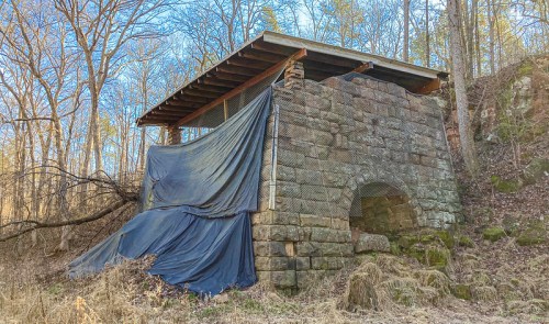 Abandoned stone structure covered with a tarp, surrounded by trees and underbrush in a wooded area.
