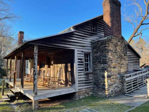 A rustic wooden cabin with a stone chimney, featuring a porch and rocking chairs, set against a clear blue sky.