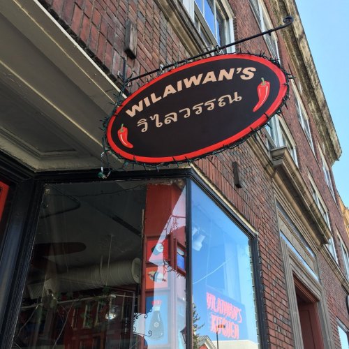 Sign for "Wilaiwan's" in Thai, hanging from a brick building, with a red and black color scheme.