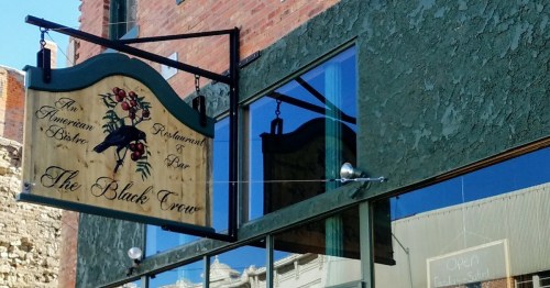 Sign for "The Black Crow" restaurant, featuring floral design, hanging from a building with large windows.